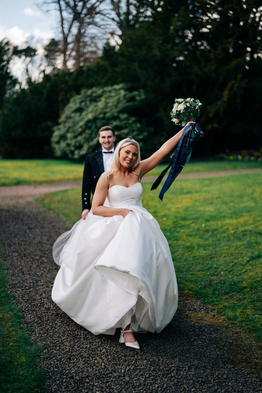 a bride wearing an ivory strapless ballgown raising a white bouquet aloft while walking on a gravel path through a green lawn. A groom in a kilt walks behind her