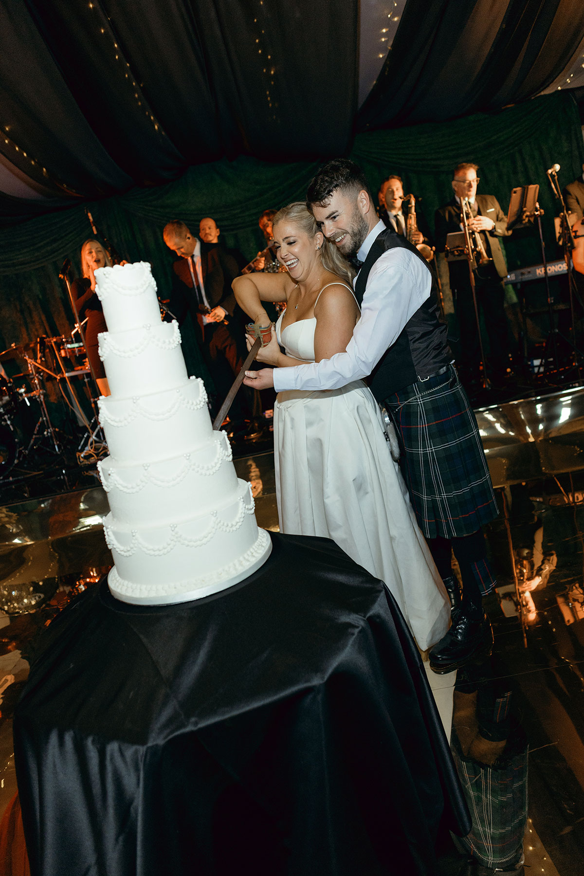 Couple cut tall white tiered wedding cake with sword during evening reception at Dundas Castle.
