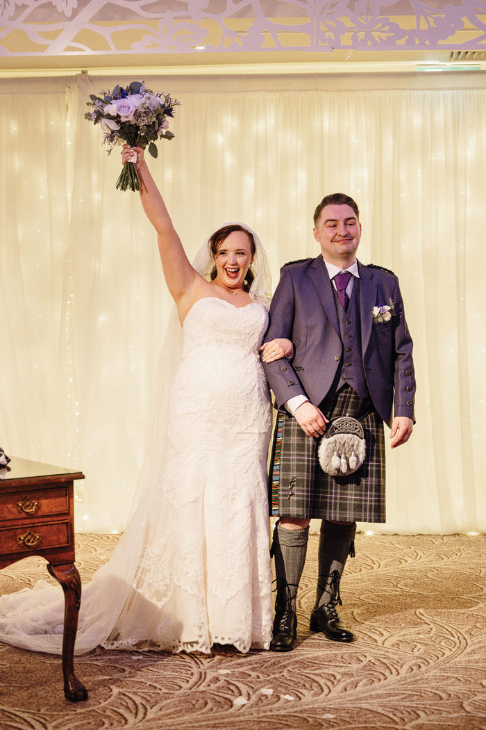 groom in full tartan outfit and bride in strapless floral wedding dress with long veil stand arm in arm, smiling as bride lifts her bouquet into the air