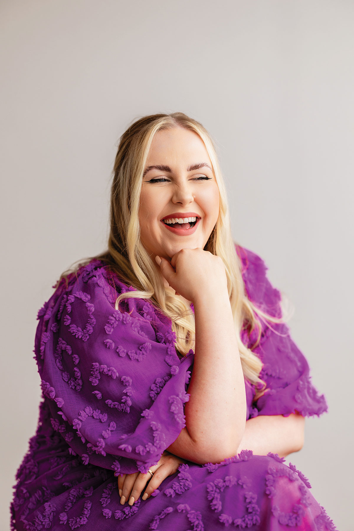 Woman with long blonde hair smiling and resting her chin on her hand, wearing a textured purple dress against a neutral studio background