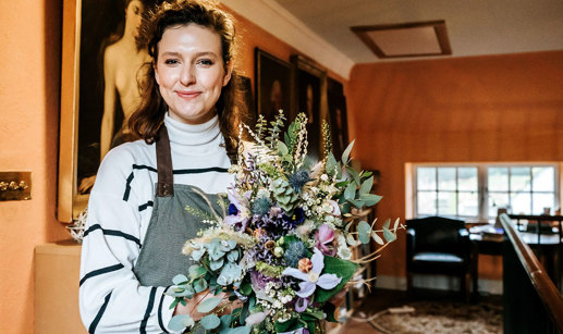 front facing woman with brown hair, a white and navy striped jumper, a green and brown apron holding a bouquet of lilac flowers and green foliage