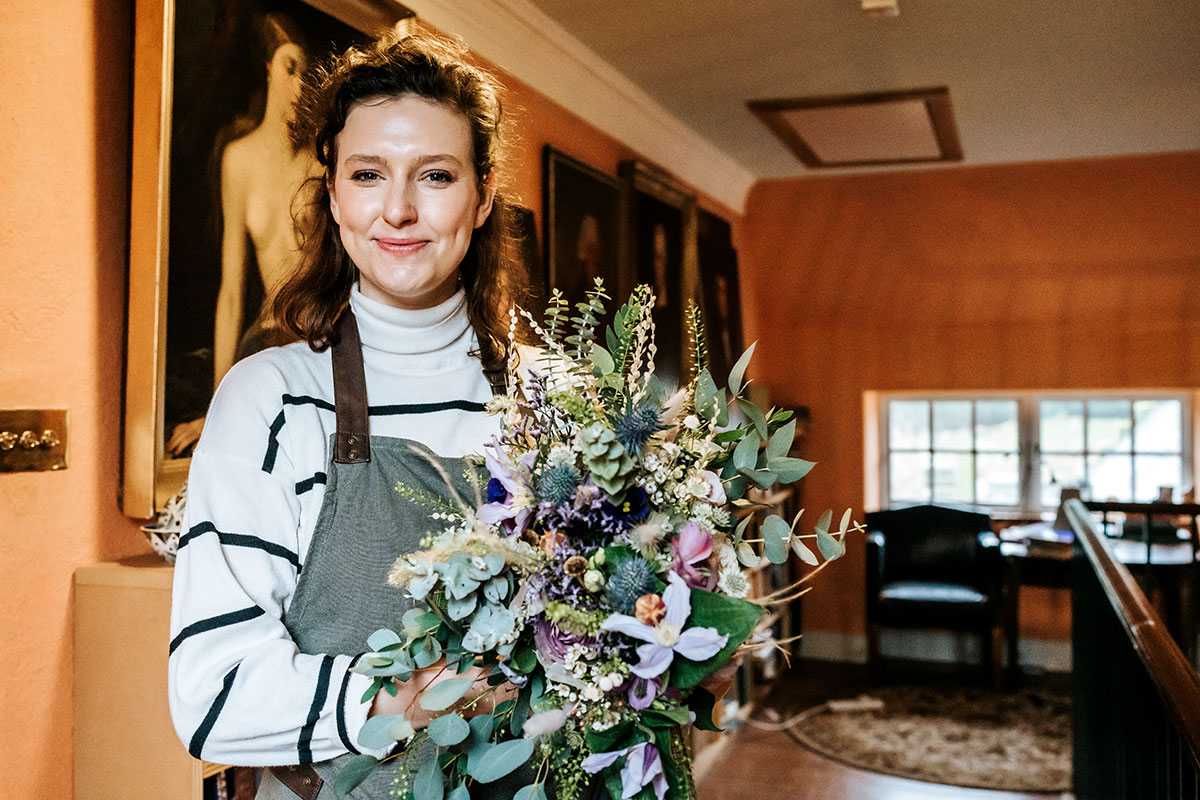 front facing woman with brown hair, a white and navy striped jumper, a green and brown apron holding a bouquet of lilac flowers and green foliage