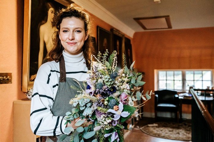 front facing woman with brown hair, a white and navy striped jumper, a green and brown apron holding a bouquet of lilac flowers and green foliage