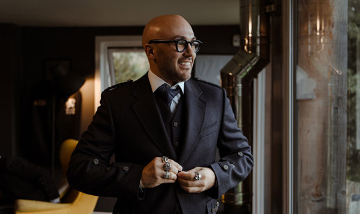 groom in navy jacket, waistcoat and tie stands looking out a large window, elbows out, and multiple large rings on show as he buttons the jacket