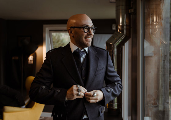 groom in navy jacket, waistcoat and tie stands looking out a large window, elbows out, and multiple large rings on show as he buttons the jacket