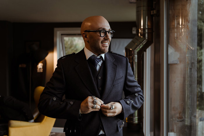 groom in navy jacket, waistcoat and tie stands looking out a large window, elbows out, and multiple large rings on show as he buttons the jacket