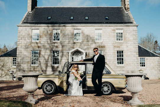 a bride and groom wearing sunglasses posing by a vintage yellow car outside Elrick House in Aberdeen