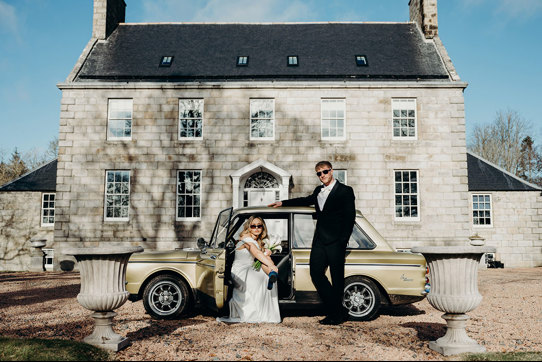 a bride and groom wearing sunglasses posing by a vintage yellow car outside Elrick House in Aberdeen