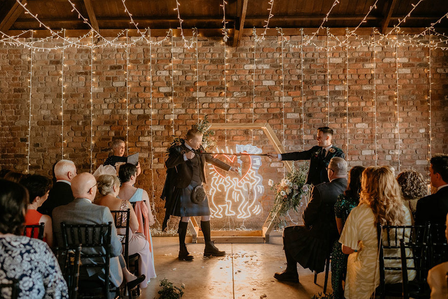 Two Grooms Handfasting With Red Brick Wall And Keith Haring Neon Sign In Background