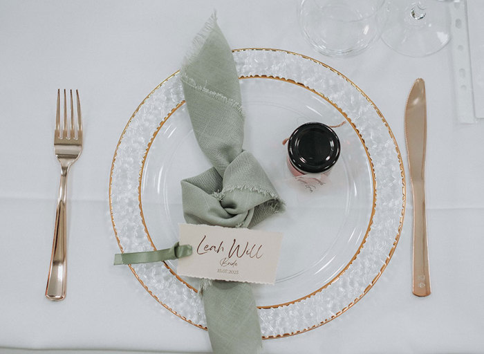 table place setting at a wedding with clear charger plate with gold rim, green knotted napkin, mini jam jar and gold cutlery