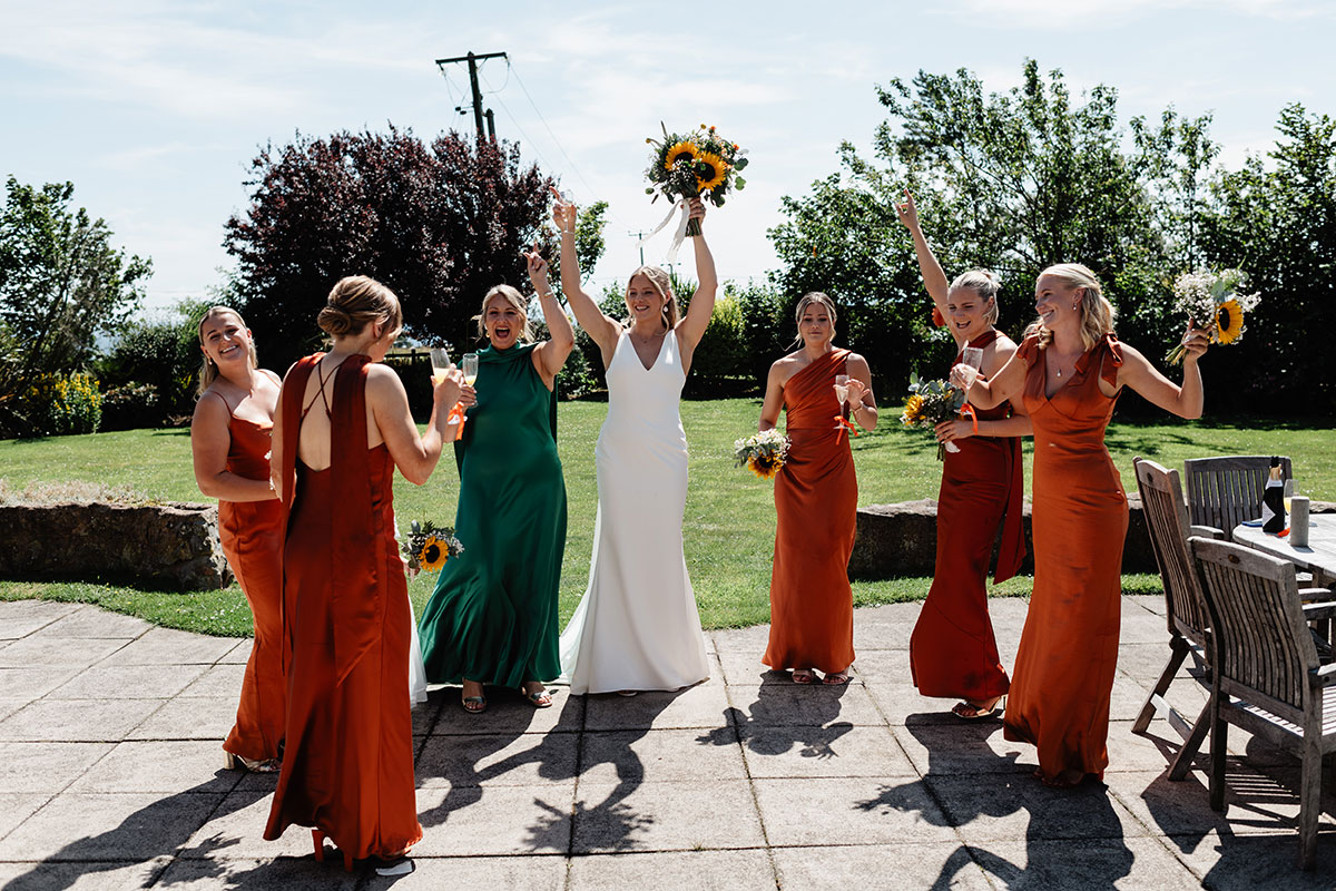 Bride and bridesmaids in rust and green dresses cheering and holding bouquets and drinks in a garden setting