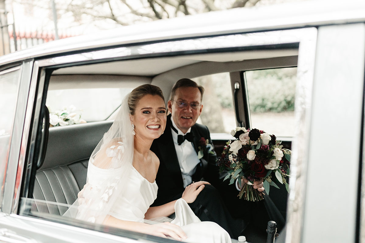Bride and her father smiling from the back seat of a classic wedding car, holding a bouquet of red and white flowers.