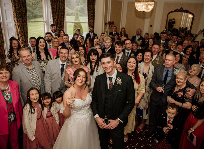 A bride and groom stand in front of their wedding guests all facing the camera and smiling