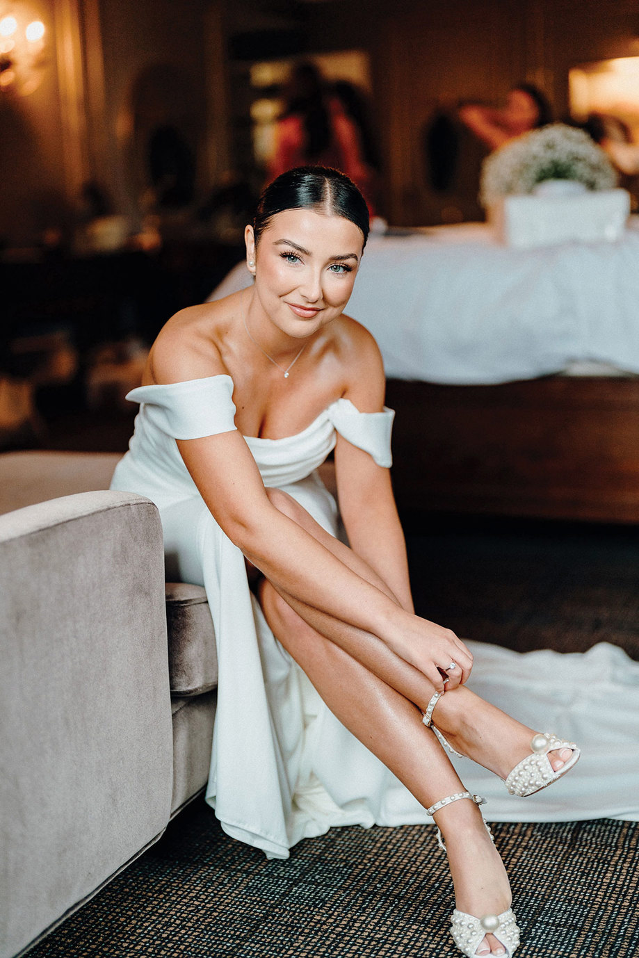 brunette bride wears slim off the shoulder strap wedding dress sitting on sofa and fixing her ankle straps on her pearl heels