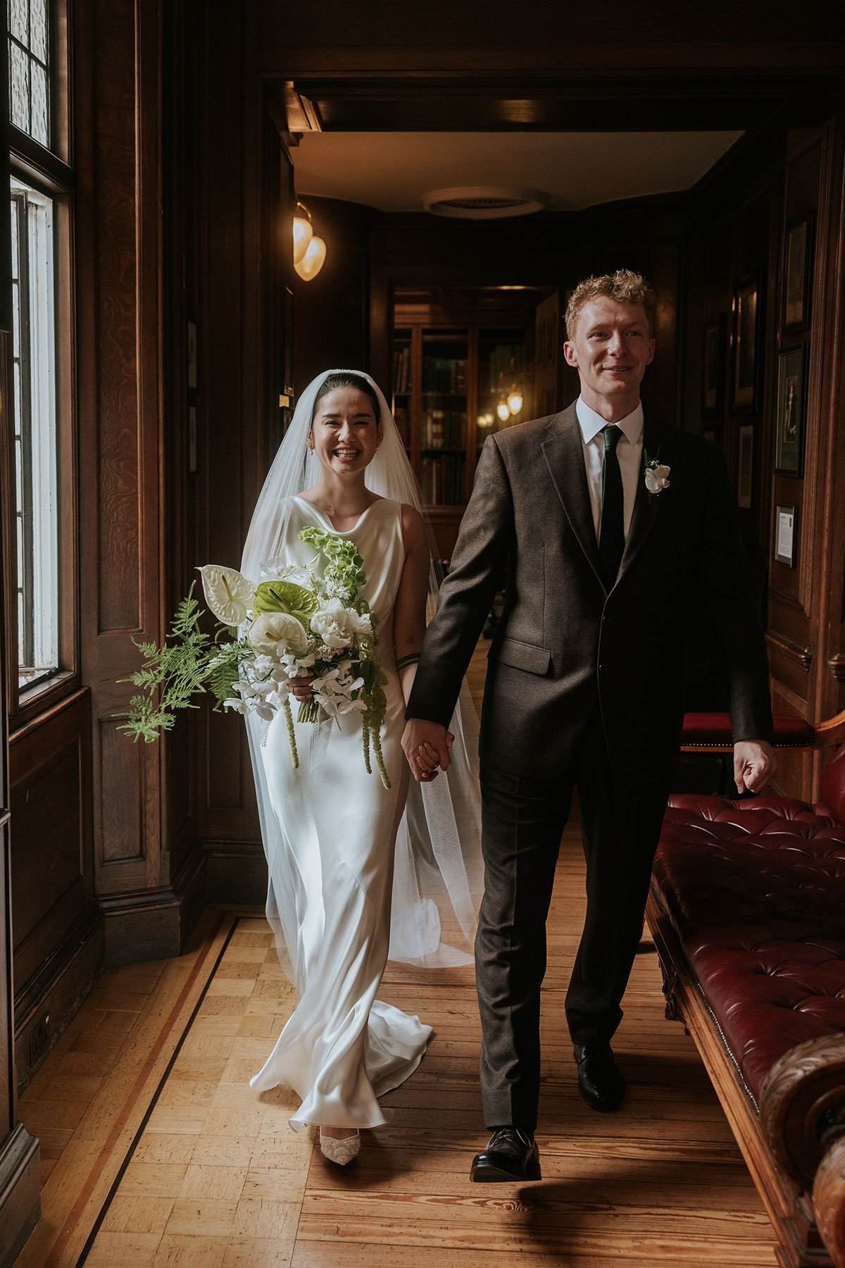 Bride and groom walking hand in hand down a corridor, bride holding bouquet.