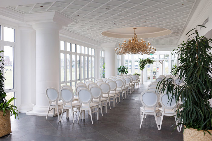 Elegant and minimalist Old Course Hotel wedding space with rows of white chairs, large windows, and a grand chandelier illuminating the room