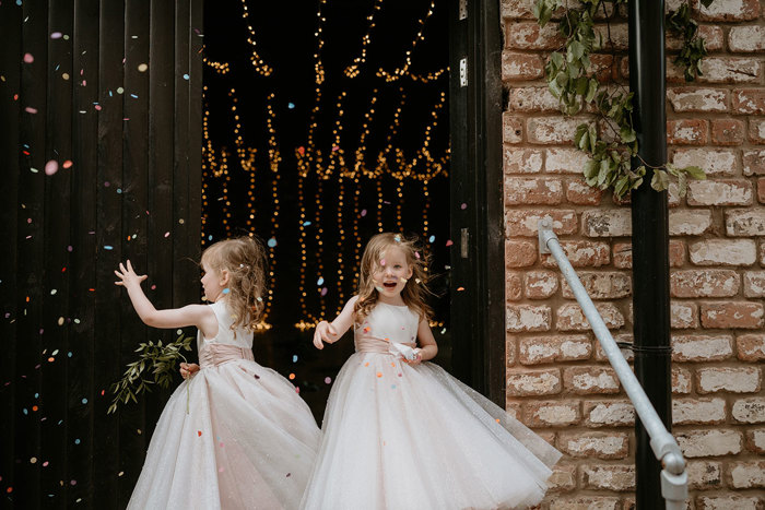 Two Flower Girls Twirling With Confetti Next To A Blonde Brick Entrance