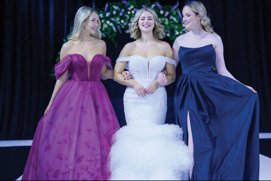 Three models smiling and walking arm in arm on the catwalk at The Scottish Wedding Show. Middle model is wearing an off-shoulder ivory gown with tulle tiered fishtail skirt. Model on right is wearing a navy bridesmaid dress. Model on left is wearing a purple off shoulder dress with floral pattern