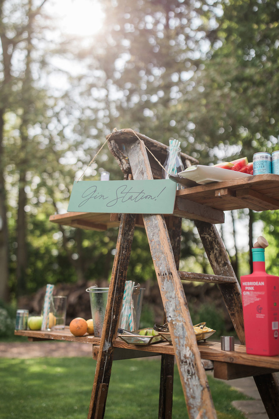 a green sign reading 'gin station' hangs from a wooden frame with shelves containing gin bottles, fruit and cans of tonic in a garden setting