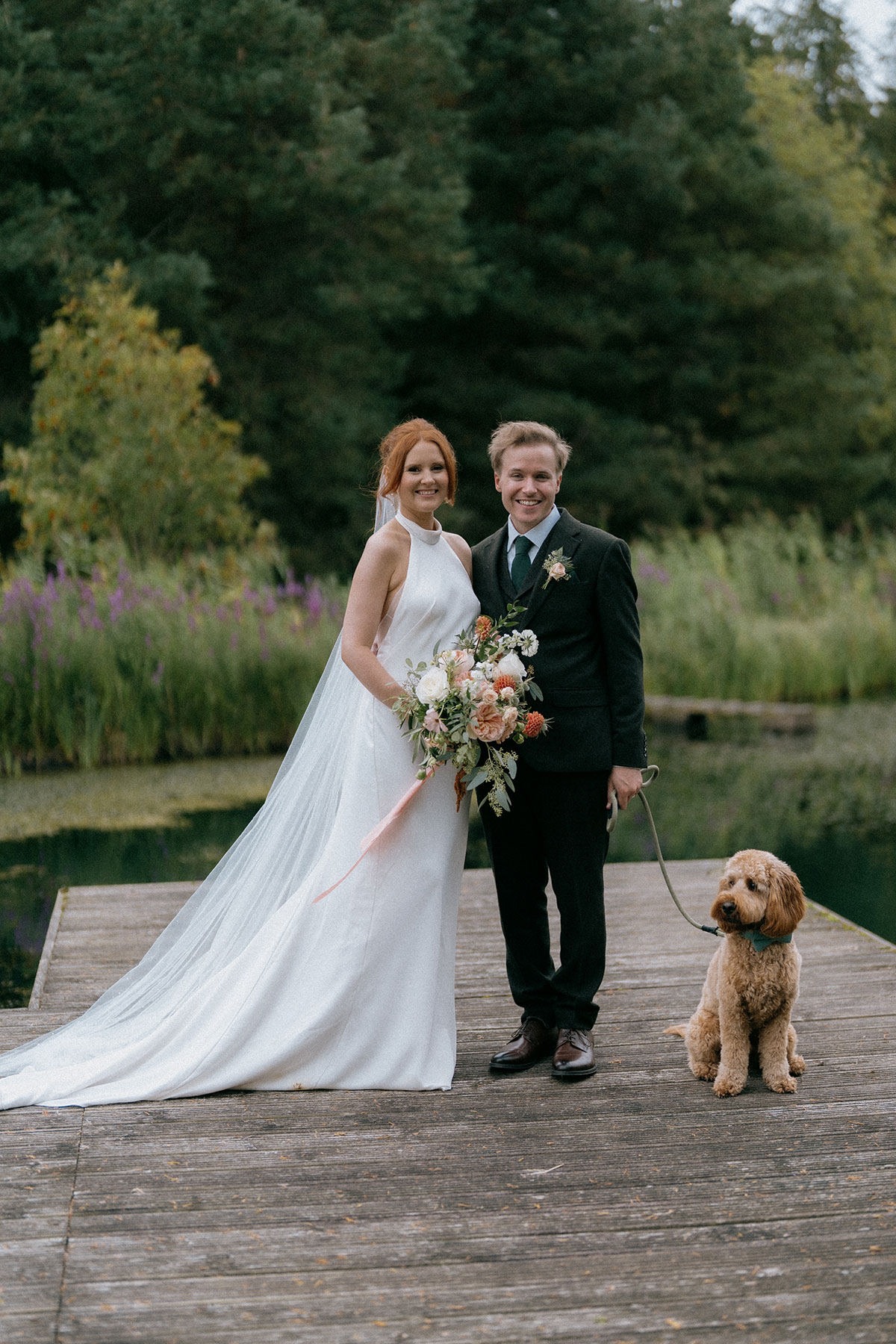 Bride and groom posing on wooden jetty by pond with their dog on lead