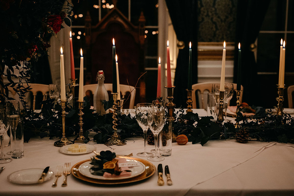 Close-up of an elegant wedding table with tall candles in brass holders, greenery, and gold-rimmed plates in a dimly lit setting