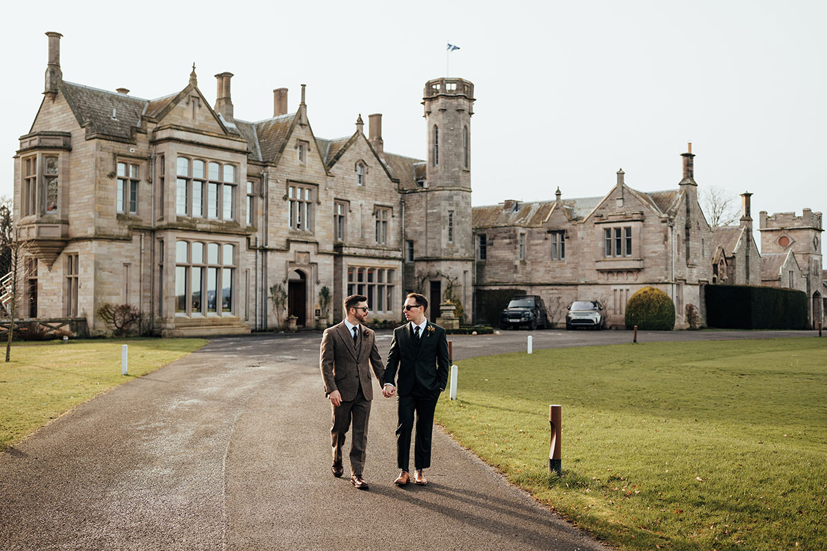 The grooms walk hand in hand along the driveway at Schloss Roxburghe