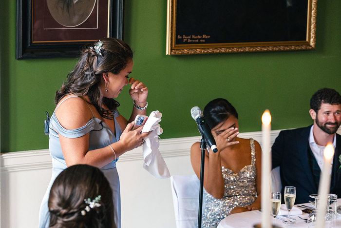 an emotional bridesmaid wearing a pale blue dress wipes away tears while looking at bride and groom seated at a table. The bride is wearing a silver sequin dress and also appears to be emotional, covering her face with one of her hands, and is comforted by a person wearing a dark blue suit