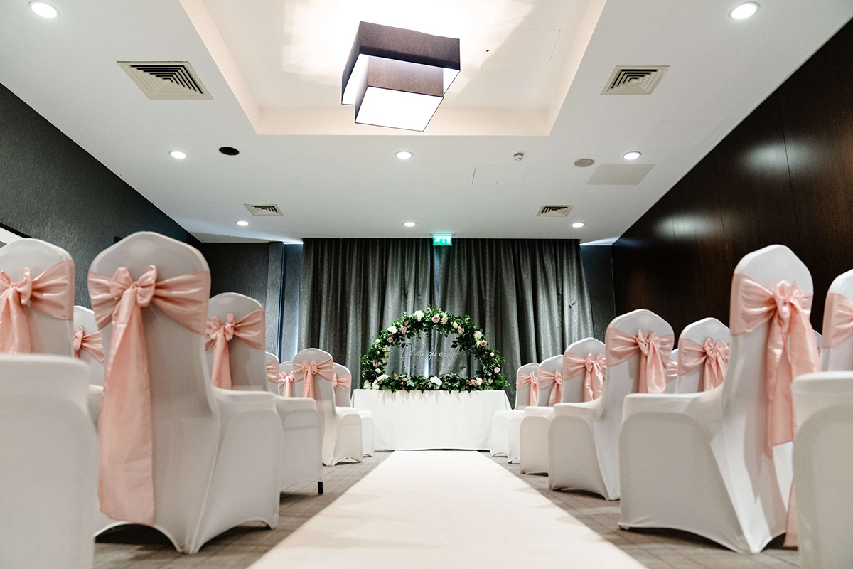 Indoor wedding ceremony room at Village Hotel Edinburgh with white chair covers, blush pink bows and a floral ceremony backdrop.