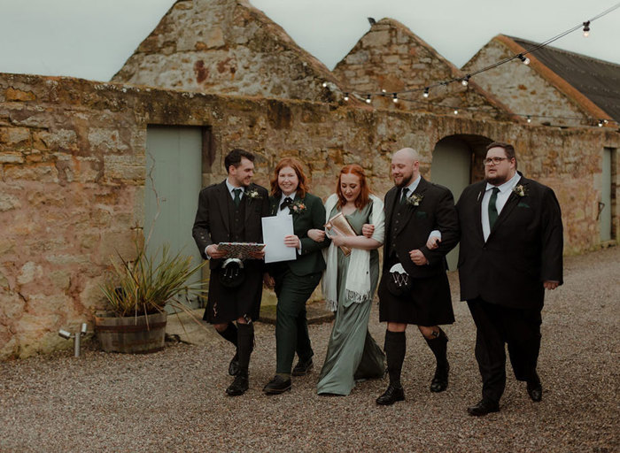 a row of five people wearing smart wedding attire walking arm and arm across gravel next to a ruined barn building. There are festoon lights strung above them 