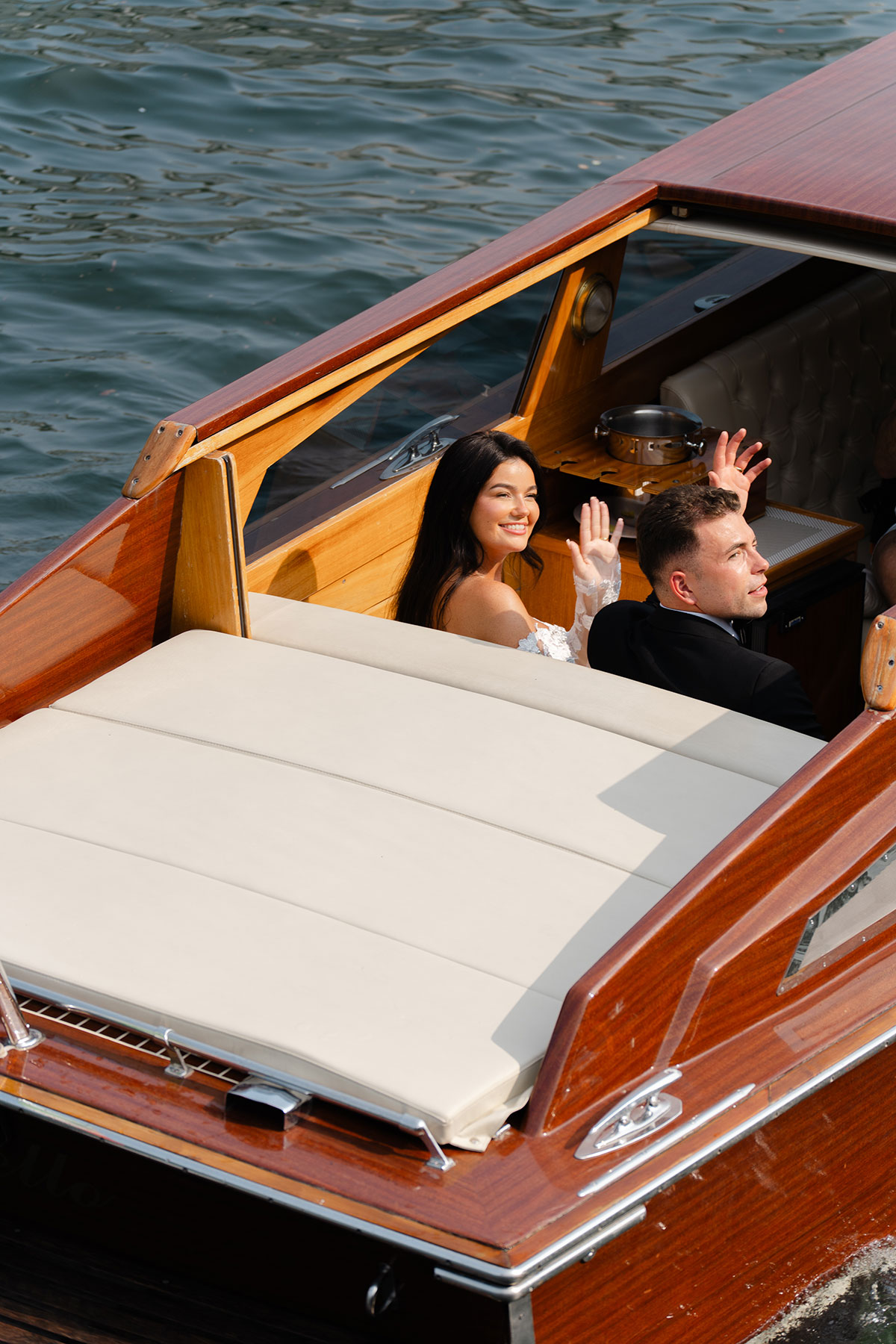 Bride and groom waving from a wooden boat on Lake Como following their Italian destination wedding