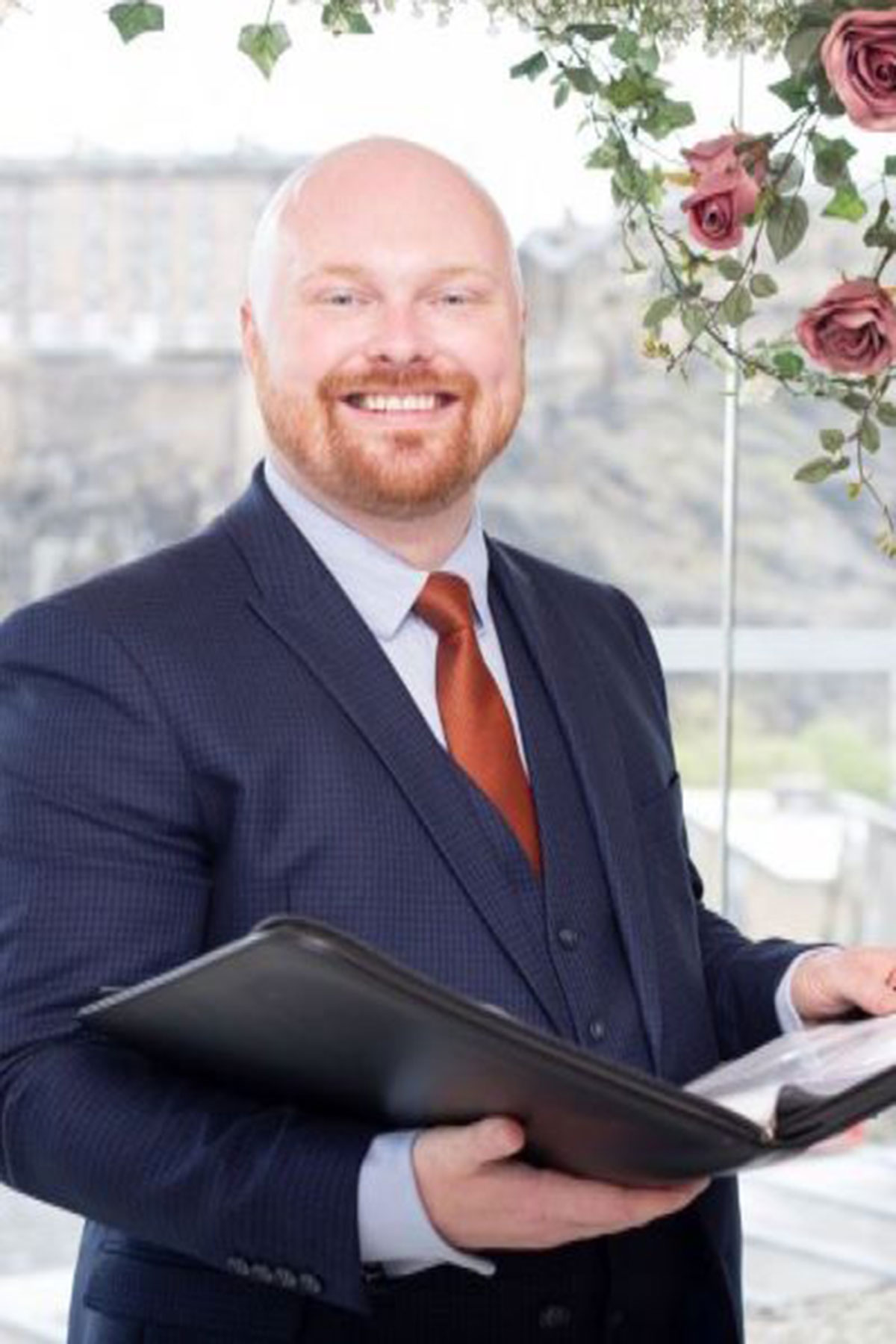 Male wedding celebrant holding ceremony folder indoors, wearing navy suit and rust tie beside floral wedding arch