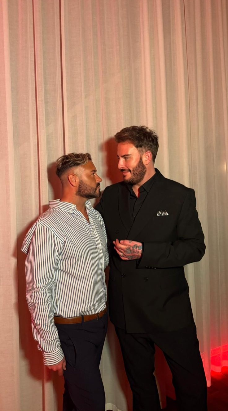 dean rowland wears a striped shirt and looks towards fiance steven wearing a black suit