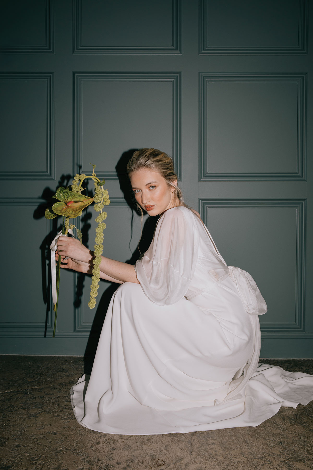 A bride in a white gown crouches down while holding a green anthurium bouquet, photographed against a dark teal panelled wall.
