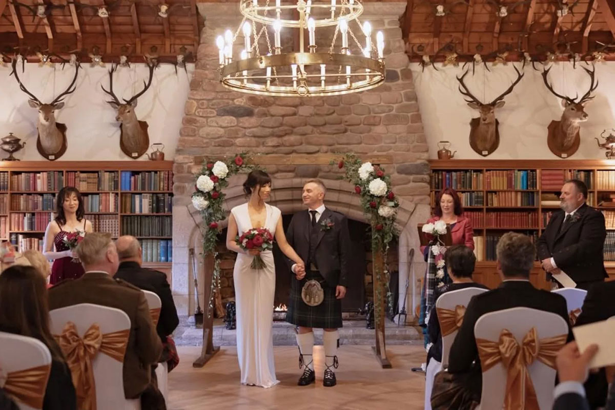 bride in white wedding dress and groom in kilt outfit hold hands at the top of aisle with floral arch and a library of books in the background