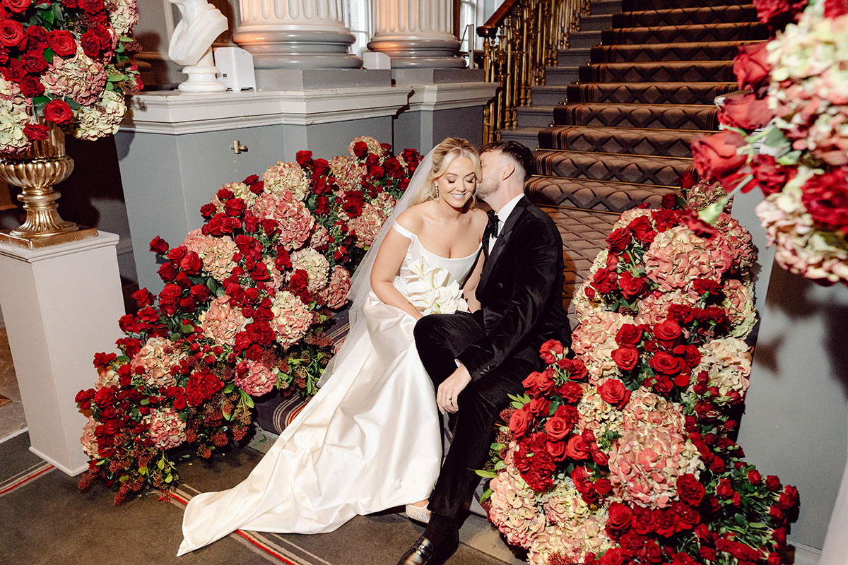 Bride and groom sitting at the bottom of the staircase at The Signet Library, surrounded by red and blush flowers