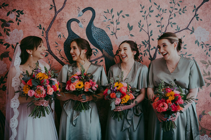 a bride and three bridesmaids holding colourful bouquets standing against a pink wall with birds and leaves painted on it at Netherbyres House