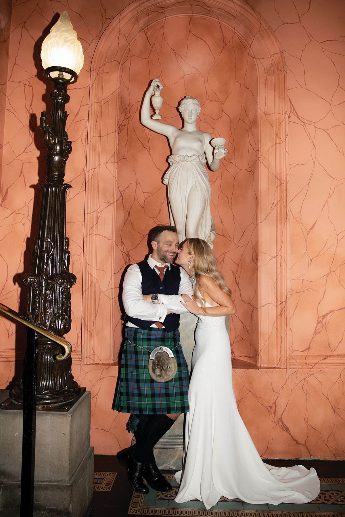 A bride and groom posing beneath a classical statue in a marble alcove at the Royal College of Physicians of Edinburgh, wearing modern bridal attire and a tartan kilt