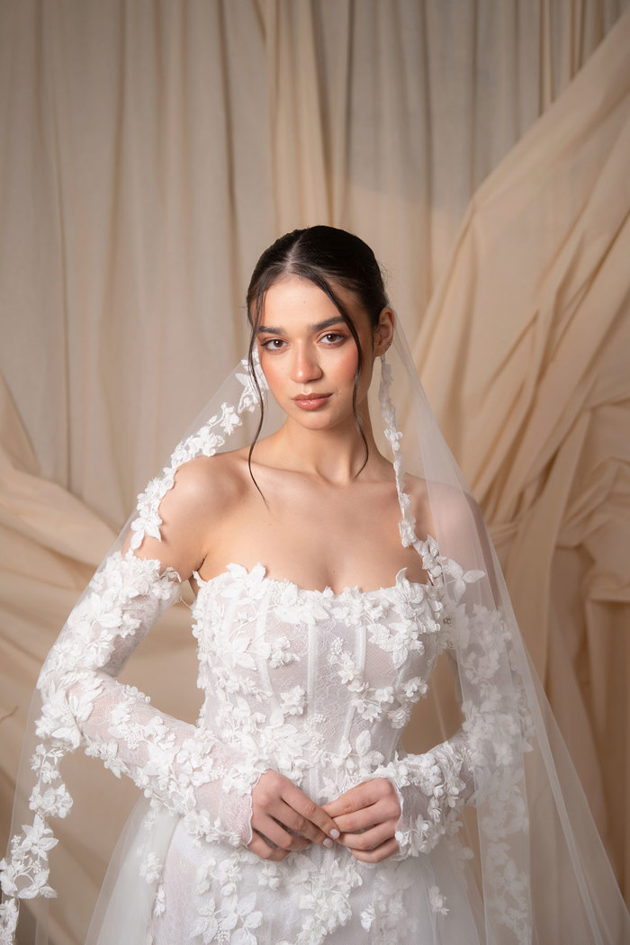 bride with tied back dark brown hair and moneypiece strands wears strapless floral applique wedding dress and matching floral border full-length veil as she stands in front of a dark cream sheet backdrop
