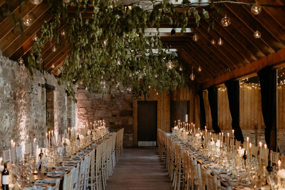 Candlelit wedding breakfast inside The Byre at Inchyra, with long tables, greenery and hanging lights.