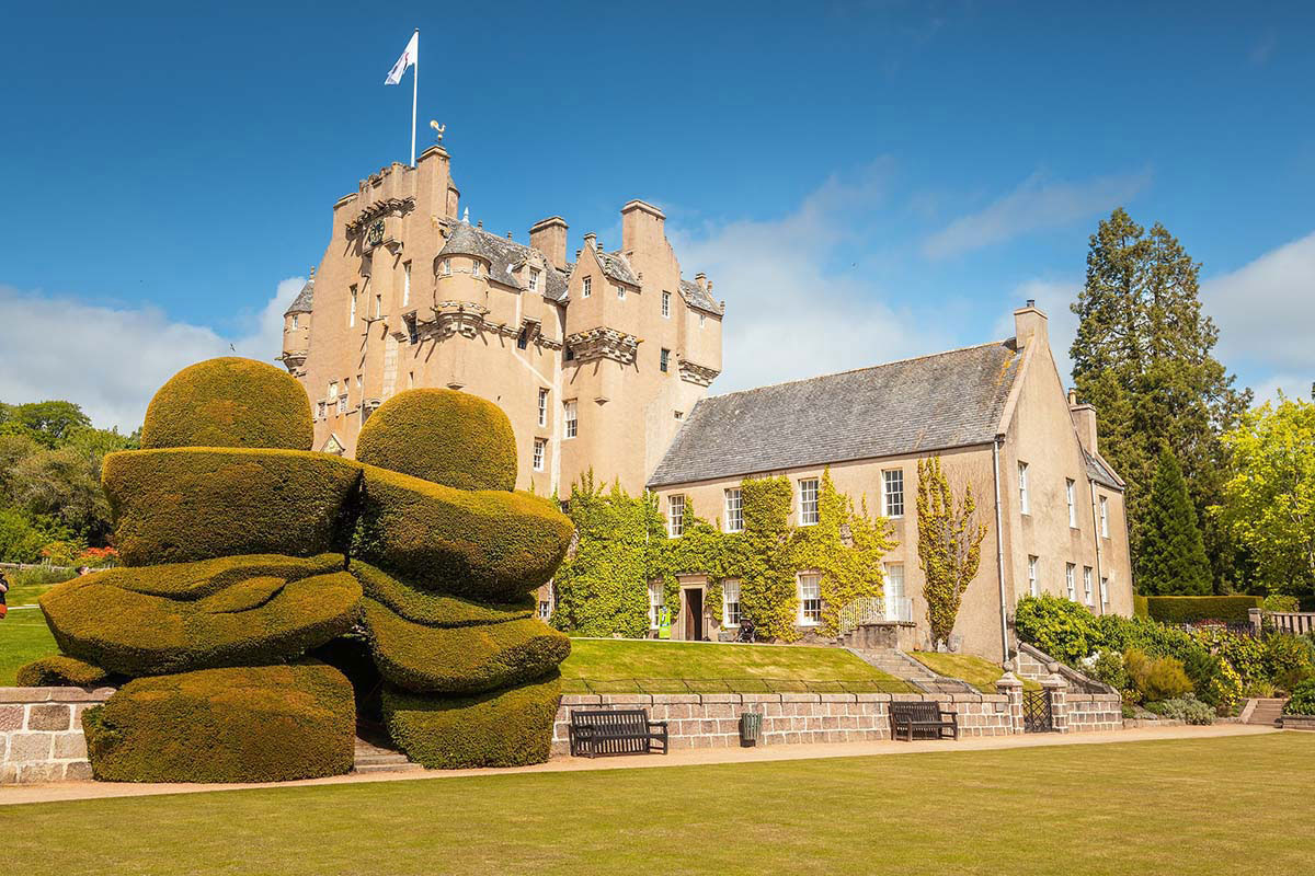 Exterior view of castle with ivy-covered walls, manicured lawns, and sculpted topiary under a blue sky