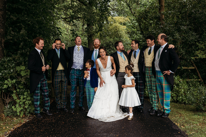 A Wedding Group Portrait With Men Wearing Tartan Trousers, A Bride Wearing A V Neck Dress And Young Children In Wedding Clothes