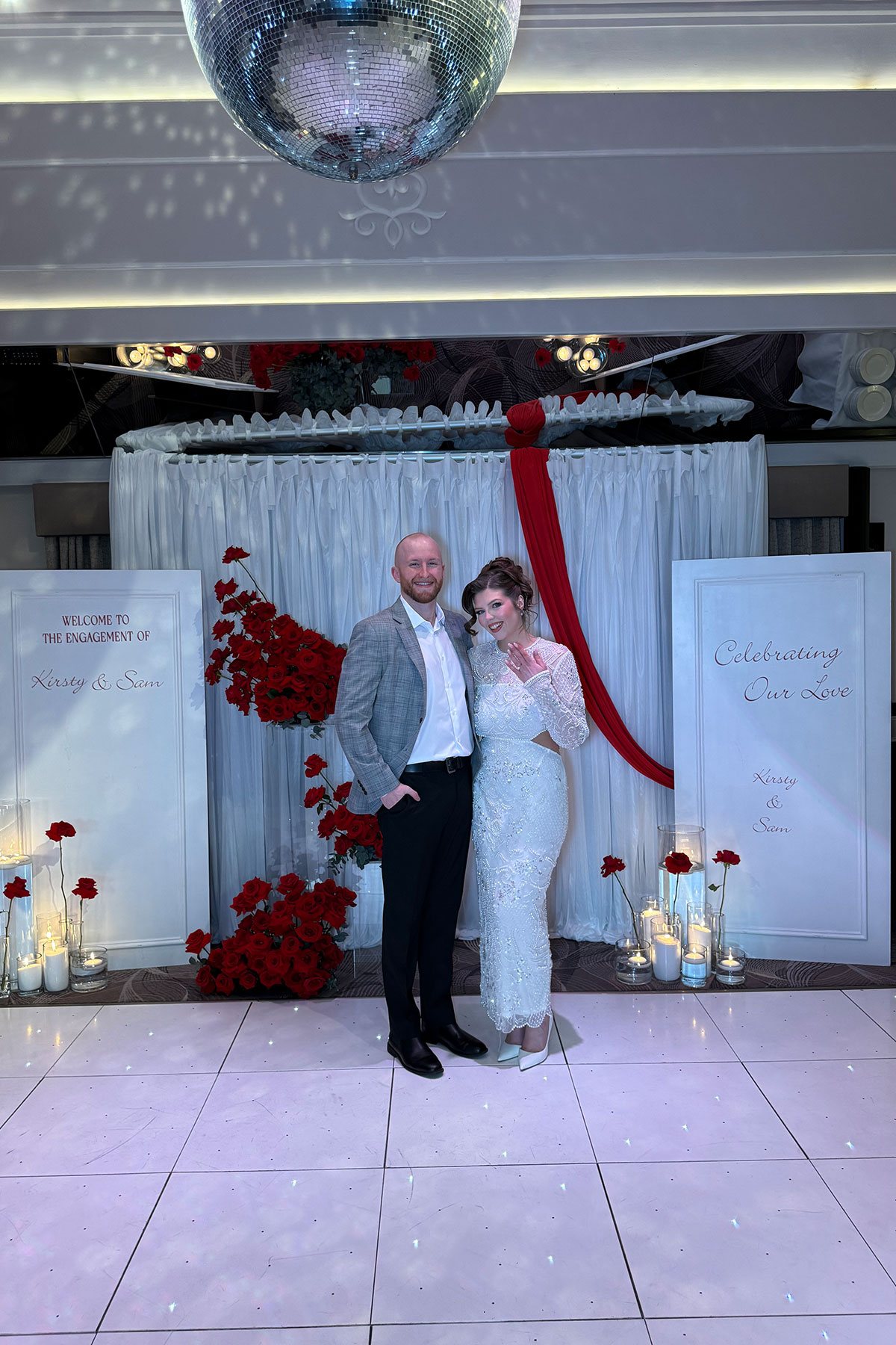 Newly engaged couple posing beside personalised red rose engagement party backdrop