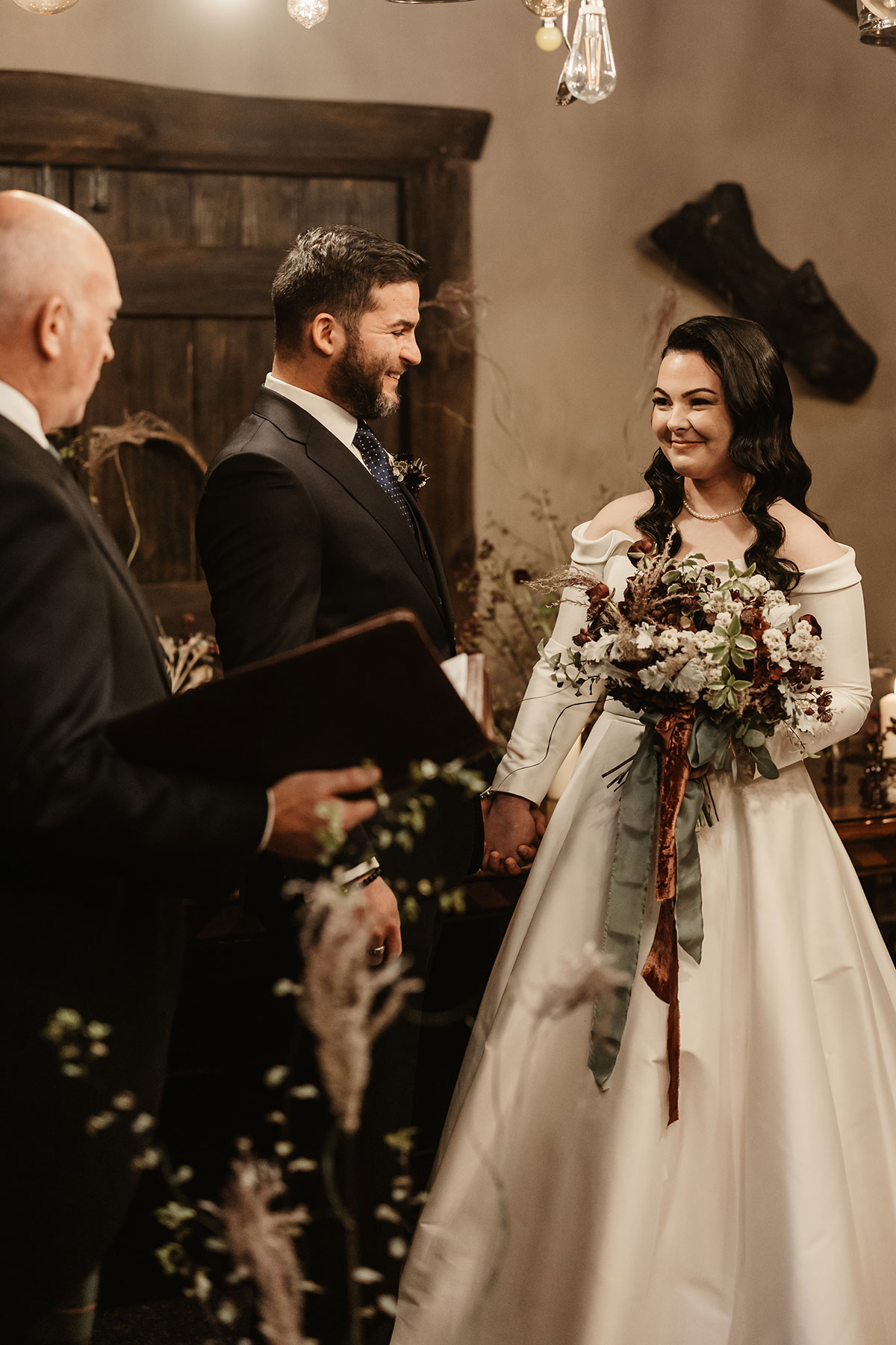 Bride and groom holding hands during the ceremony, smiling at each other beneath warm lighting
