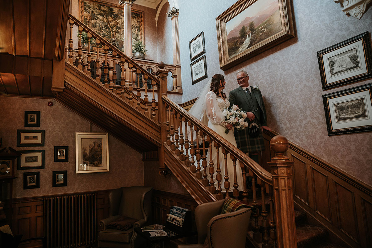 Bride holding bouquet while walking downstairs with her father before wedding ceremony