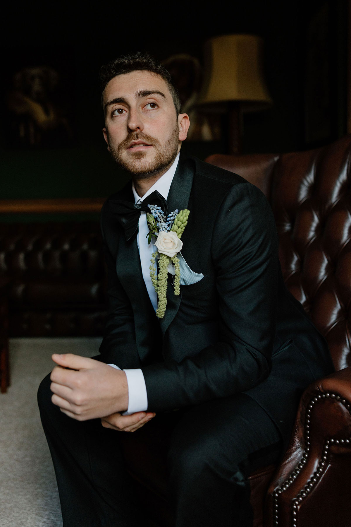 Groom in a black tuxedo seated in a brown leather armchair