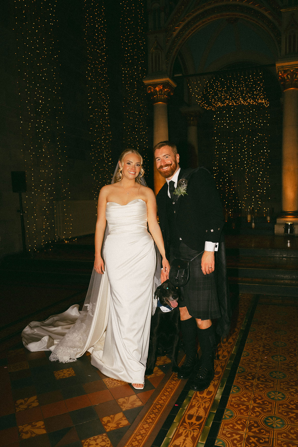bride and groom with dog posing inside candlelit wedding venue with fairy lights