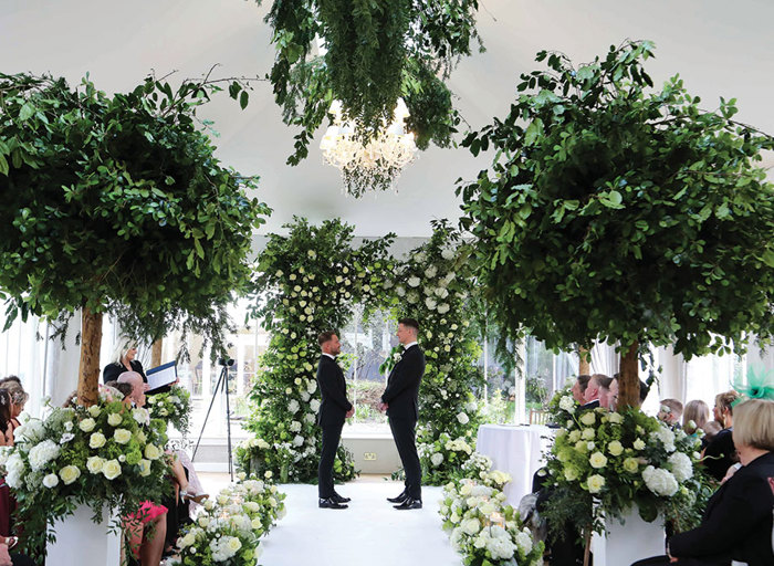 two grooms stand at the top of a white aisle during their wedding ceremony in the conservatory at Carlowrie Castle. There is a large green and white floral rectangular arch behind them, green trees in white pots either side of the aisle and lots of green and white floral arrangements on the floor. Wedding guests are seated either side of the aisle and an officiant stands at the left hand side holding an open binder