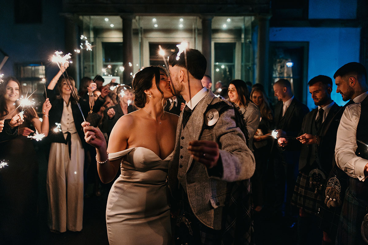 bride in off the shoulder wedding dress and groom in tweed kilt outfit kiss outdoors holding sparklers and surrounded by friends and family in the dark, also holding sparklers and smiling