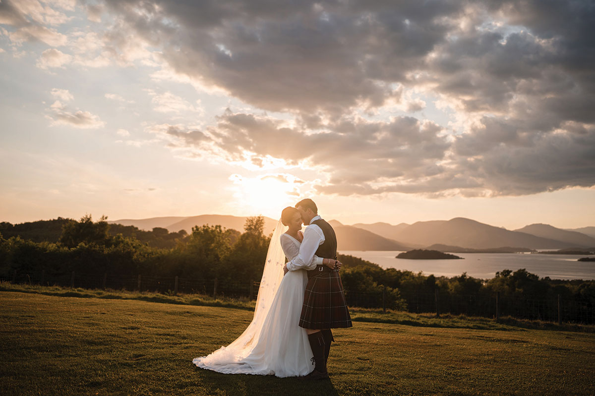 bride in flowy wedding dress and floor-length veil and groom in red kilt outfit embrace on grass with lochs, hills and a sunset sky in the background