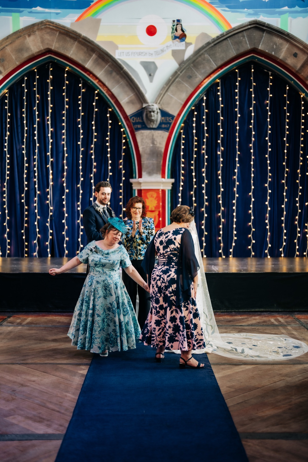 Family members take part in Robyn and James's wedding ceremony at Òran Mór in Glasgow, beneath colourful murals and fairy lights.